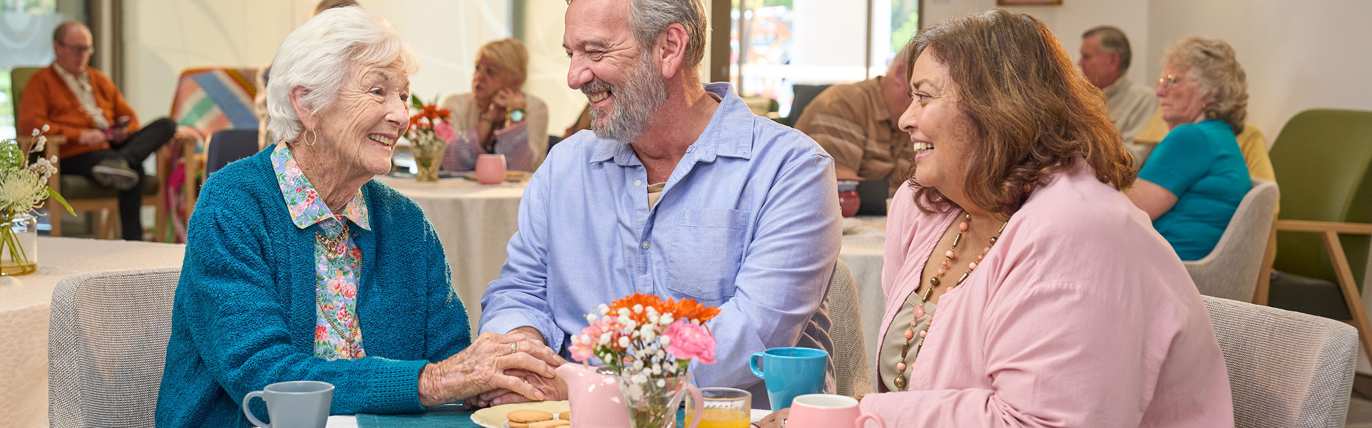 An elderly woman sitting in a dinning room with a man and women, holding the mans hand.